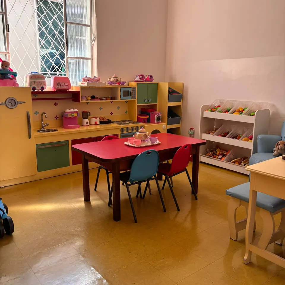 O cantinho da casinha, com cozinha de brinquedo, mesa vermelha com um jogo de chá e cadeirinhas